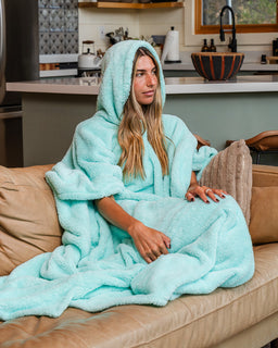 A woman sits on a tan couch wrapped in a fluffy hooded bathrobe in a bright kitchen setting. The robe is a light aqua/mint color and has a plush, oversized texture.