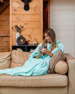 A woman sits on a tan leather couch wrapped in a fluffy light aqua blanket, holding a mug in a cozy cabin-style living room with wood-paneled walls and decor.