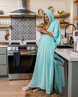 A woman in a cozy kitchen wearing a plush, hooded bathrobe in light aqua/mint green. She is smiling and holding a mug, standing in front of a stove and tiled backsplash.