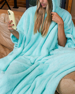 A woman sitting on a tan couch wrapped in a plush hooded blanket, holding a cream-colored smartphone.