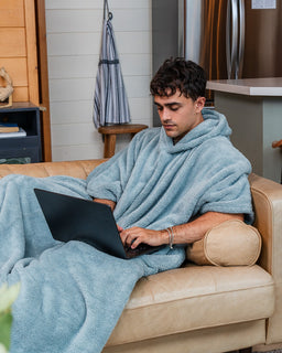 A man sits on a beige leather couch wrapped in a light blue fleece blanket hood, using a black laptop in a modern kitchen setting.