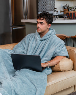 A man sits on a beige leather couch in a modern kitchen setting, wrapped in a light blue plush hooded blanket and using a black laptop.