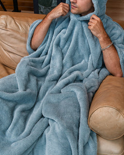 A man reclining on a tan leather couch, wrapped in a plush hooded blanket that covers his head and body. The blanket is a dusty light blue color, and the background shows a cozy indoor setting.