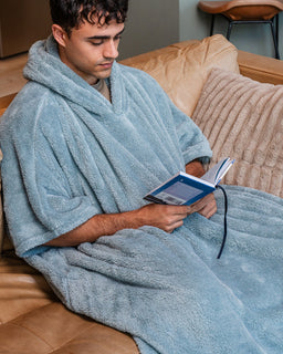 A man sitting on a tan leather couch wrapped in a plush light blue hooded blanket or robe, reading a book.