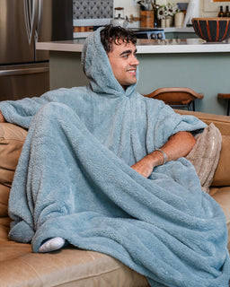 A man sitting on a tan leather couch wrapped in a light blue hooded fleece blanket, with a kitchen visible in the background.