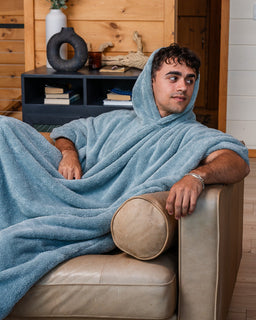 A man reclining on a beige leather couch, wrapped in a light blue hooded fleece blanket. The background shows a cozy room with wood-paneled walls and a dark shelf.