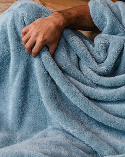 A close-up of a person wrapped in a soft, plush light blue blanket, showing their hand and arm as they pull the fabric.