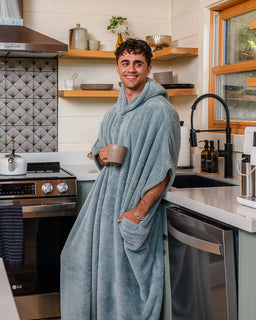 A man wearing a plush hooded bathrobe in a modern kitchen, holding a beige mug. The robe is a dusty blue-gray color with a hood and front pocket.