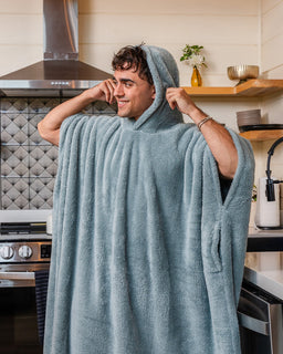 A man wearing a plush, hooded bathrobe in a modern kitchen setting. The robe is a dusty slate blue color and features a hood, long sleeves, and side pockets.