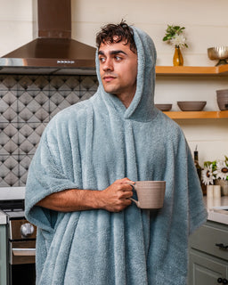 A man wearing a light blue hooded bathrobe stands in a kitchen holding a beige ceramic mug.