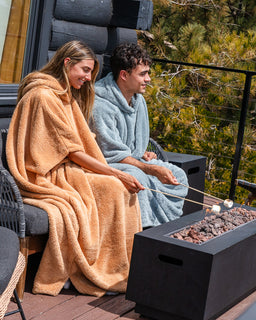A man and a woman sitting on a wooden deck next to a fire pit, wrapped in plush hooded blankets. The woman's blanket is a warm camel tan, and the man's is a light dusty blue.