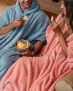 A cozy lifestyle scene of a man and a woman sitting together on a couch, wrapped in plush fleece blankets in dusty blue and dusty pink, respectively. The man holds a bowl of potato chips while both hold chips in their hands, set against a warm wooden floor background.