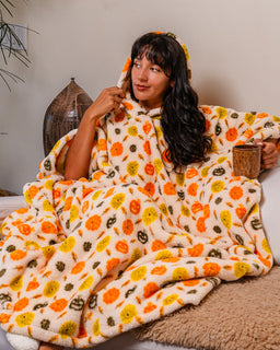 A woman sits wrapped in a cozy hooded fleece blanket with a colorful pumpkin print, holding a mug in a warm indoor setting.