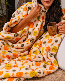 A cozy cream-colored plush blanket with a vibrant orange, yellow, and olive green floral pattern, wrapped around a person holding a wooden mug.