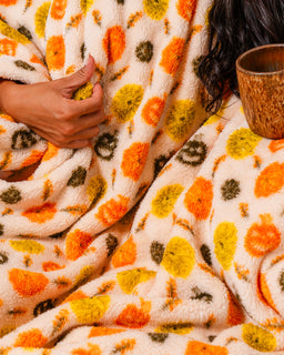 A cozy close-up of a person wrapped in a plush cream throw blanket with a colorful pattern of orange, yellow, and olive green abstract shapes. A wooden mug is visible on the right side.