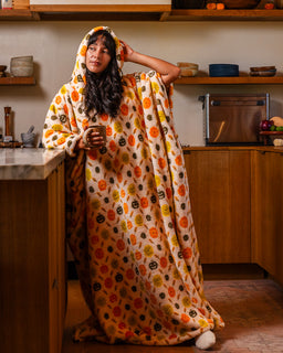 A woman stands in a warm kitchen wearing a hooded blanket or sleeping bag with an orange, black, and yellow pumpkin pattern. She holds a mug and looks directly at the camera.