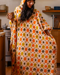 A woman in a cozy kitchen wearing a plush hooded blanket with an orange, yellow, and brown pumpkin pattern, holding a mug.