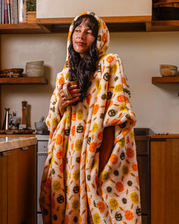 A woman wearing a hooded blanket wrap with a pumpkin pattern, standing in a cozy kitchen setting.