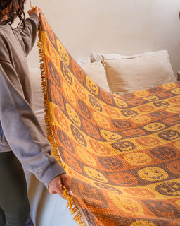 A person holds up a large woven blanket or throw with a repeating pumpkin face pattern in orange, yellow, and brown, with fringed edges, in a bedroom setting.