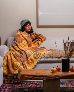 A cozy living room scene with a woman wrapped in a warm yellow and orange patterned blanket while sitting on a light gray sofa. The blanket features a repeating jack-o'-lantern design.