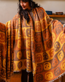 A woman wearing a large woven blanket or shawl with a patchwork pattern of orange, yellow, and brown pumpkin faces, shown in a warm indoor setting.