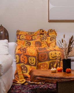 A cozy living room scene featuring a thick throw blanket draped over a white armchair. The blanket has a warm autumnal design with a repeating pattern of jack-o'-lantern faces in orange, brown, and yellow.