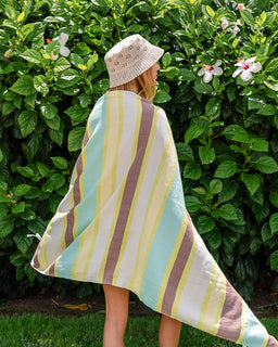 A woman standing outdoors in front of a green leafy hedge, wrapped in a large striped beach towel. The fabric has vertical stripes in pale yellow, mint green, dusty pink, and cream.