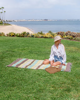 A woman sits cross-legged on a striped beach towel on a green lawn, with a beach, ocean, and palm trees in the background. A woven basket bag rests beside her.