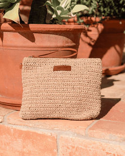 Beige woven clutch bag with a brand label on a stone surface with potted plants in the background