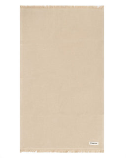 A rectangular beige hadn towel with fringed edges, displayed flat against a white background. A small white brand label is visible in the bottom right corner.