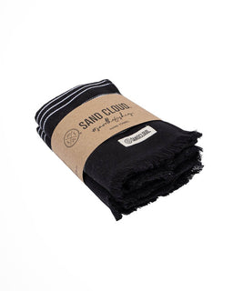 A folded black cotton towel with white stripes on one end and fringed edges, wrapped in a brown kraft paper band labeled 'SAND CLOUD'