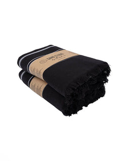 Two folded black fringed towels stacked on a white background, each wrapped with a tan paper band
