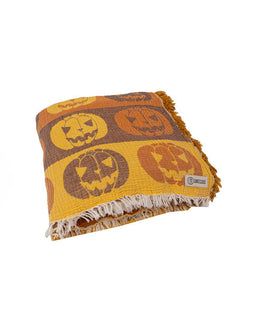 A folded woven throw blanket featuring a pumpkin pattern in orange, brown, and yellow, with cream fringe on a white background.