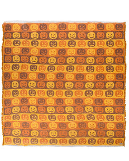 A rectangular woven textile with fringe on two short ends, featuring a repeating pattern of carved jack-o'-lantern pumpkins in orange, yellow, and brown on a warm tan background.