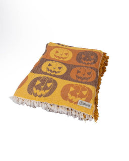 A woven throw blanket with a Halloween pumpkin pattern, featuring orange and brown jack-o'-lantern motifs on a mustard yellow background, finished with white fringe on all sides.