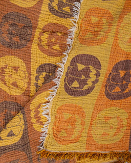 A close-up of a textured cotton blanket with a Halloween pattern, featuring orange and yellow jack-o'-lantern motifs on a warm mustard yellow background, finished with white fringed edges.