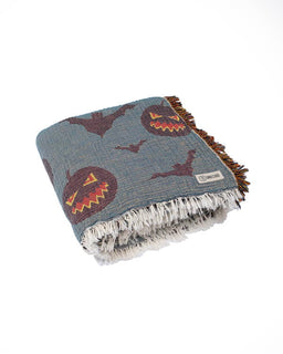A folded woven throw blanket with a slate blue-gray base, featuring a pattern of jack-o'-lanterns and bats in dark red, orange, and yellow, finished with white and brown fringe.