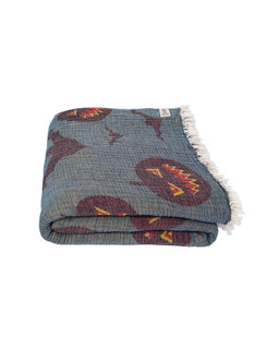 A folded woven throw blanket with a textured slate blue base, featuring a pattern of dark red, yellow, and orange abstract shapes, finished with white fringe on one end.