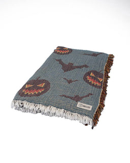 A woven throw blanket with a muted blue-gray base, featuring a pattern of jack-o'-lanterns, bats, and small birds, finished with white and brown fringe.