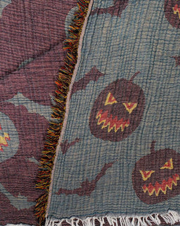 Close-up of a woven textile with a Halloween pattern, featuring jack-o'-lantern faces, bats, and colorful fringe along the edge.