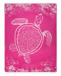 A rectangular pink towel or blanket with fringe on the top and bottom edges, featuring a large white sea turtle design with intricate circular and geometric patterns inside its shell and flippers. The background has a distressed, speckled white pattern around the edges.