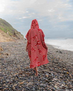 Person wearing a red and white patterned poncho on a pebbly beach with ocean and sky in the background