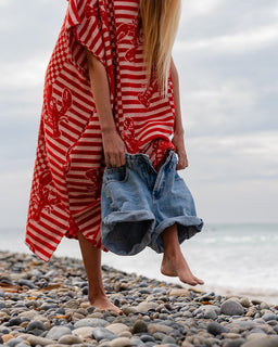 Person wearing a red and white patterned poncho and denim shorts on a pebbly beach.