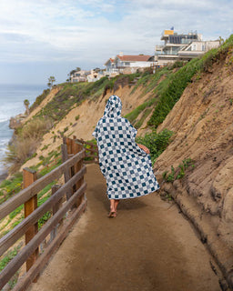 Person wearing a checkered poncho walking along a coastal path with houses on a cliff in the background.