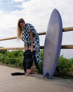 Person with a checkered towel and wetsuit standing next to a purple surfboard on a wooden deck.