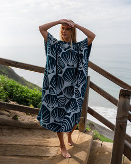 Person wearing a blue and white shell patterned towel by a coastal area.