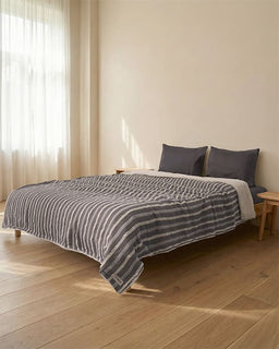 Bed with striped bedding and gray pillows in a room with wooden flooring and light-colored walls.