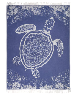 A rectangular woven textile with fringe on the top and bottom edges, featuring a large white sea turtle design on a medium blue background with distressed white speckling.