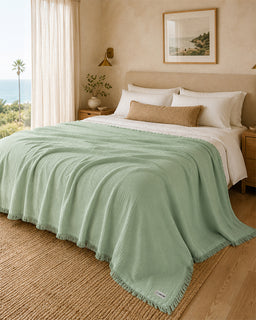 A sage green textured throw blanket with fringe trim draped over a bed in a warm, modern bedroom setting.