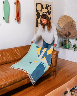 A woman in a white long-sleeve top and blue jeans holds up a woven throw blanket with a geometric pattern in light blue, navy, yellow, and white, draped over a brown leather sofa in a modern living room.